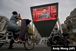 A woman on her electric-powered scooter films a large video screen outside a shopping mall showing Chinese President Xi Jinping speaking during an event to commemorate the 100th anniversary of China's Communist Party at Tiananmen Square in Beijing on July 1, 2021.
