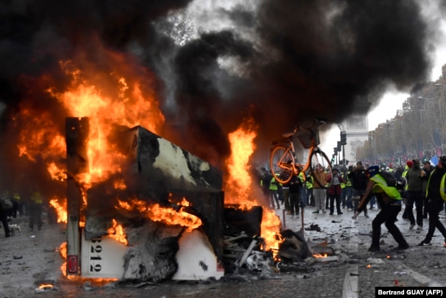 FRANCE -- A man throws a bike in a burning truck during a protest of Yellow vests (Gilets jaunes) against rising oil prices and living costs near the Arc of Triomphe on the Champs Elysees in Paris, on November 24, 2018.